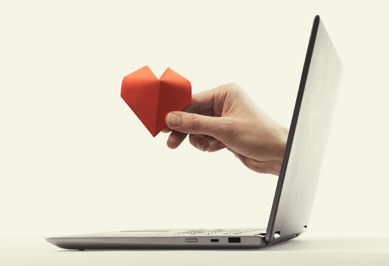 A hand emerges from a laptop screen holding a red paper heart, symbolizing online connection or support for virtual charities.