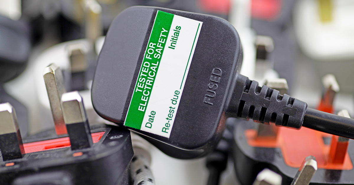 Close-up of a black electrical plug with a green sticker reading "PAT Testing." Other plugs blurred in the background.