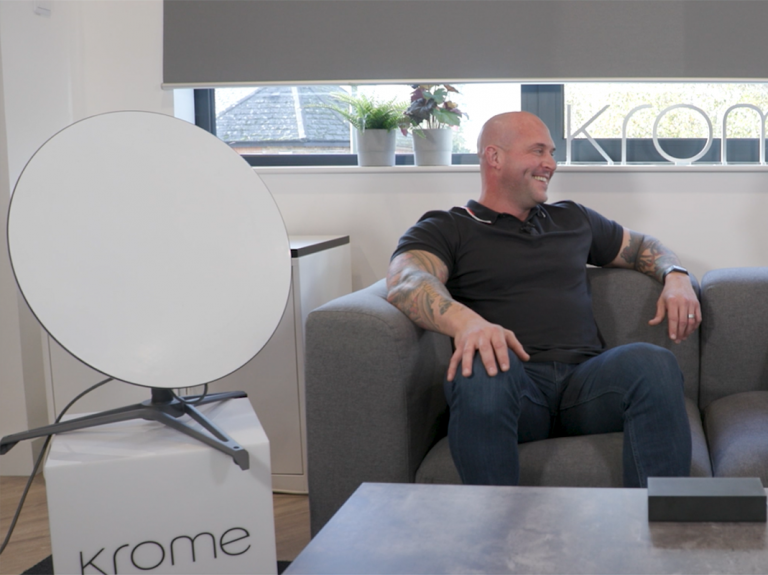 A bald man with tattoos smiles while sitting on a couch, next to a large, round Starlink Satellite light in an indoor setting.