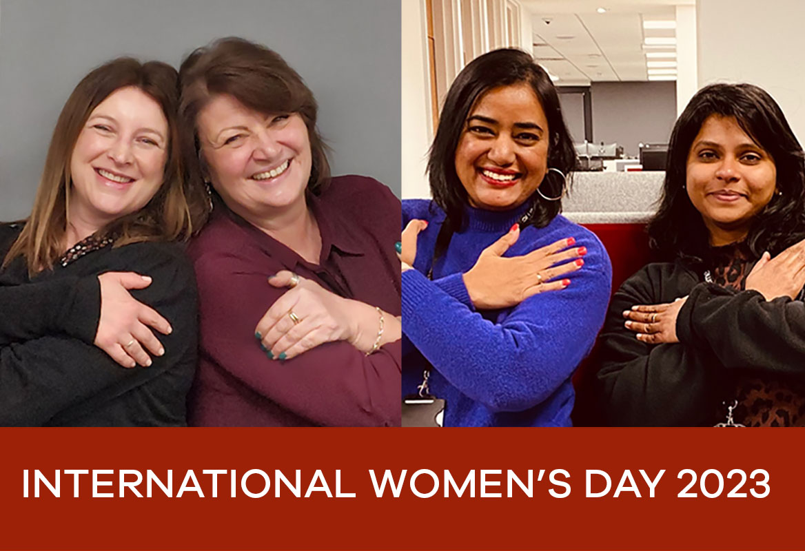 Four women smiling and embracing, celebrating International Women's Day 2023, against office backgrounds.