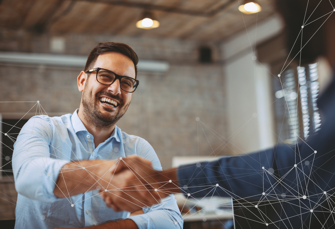 Smiling man in glasses shaking hands across a table in an office during an M&A IT integration discussion, with digital network graphics overlaid.