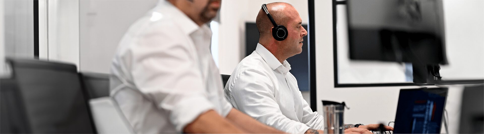 Two men in a modern office, one wearing headphones and working on a computer, focused on their tasks.