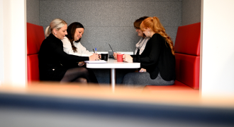 Three women sit around a small table in a modern office booth, focusing on documents and a laptop while having a discussion.