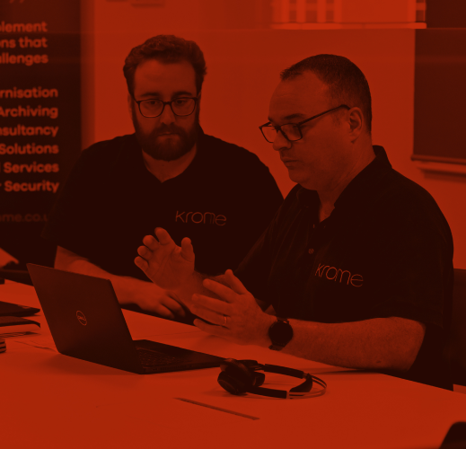 Two men in black t-shirts with "krome" logo working on a laptop at a table in an office setting.