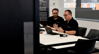 Two men in black polo shirts working together on a laptop in a modern office with a whiteboard in the background.