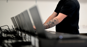 A person with a tattooed arm setting up multiple computer monitors in an office.