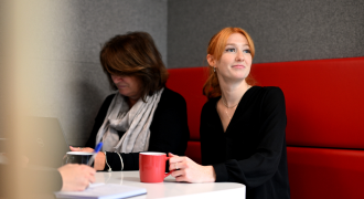 Two women sitting at a table in a modern office, one using a laptop and the other looking towards the camera.