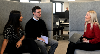 Three colleagues conversing in an office setting, two seated on a couch and one on a chair, with laptops and notebooks.