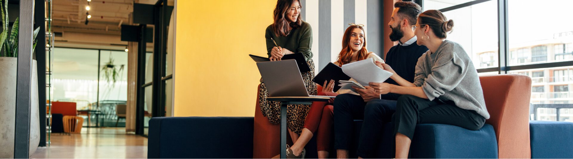 Four colleagues casually discussing Microsoft Cloud Solutions with documents and a laptop in a modern office lounge area.