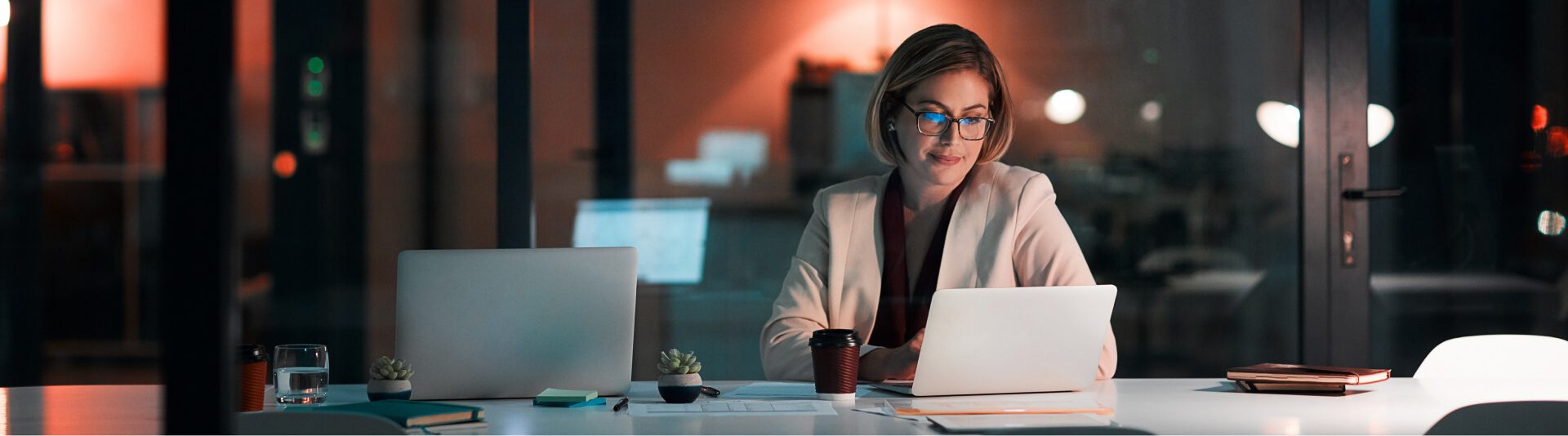 A woman works on a laptop at a desk in a dimly lit office at night, surrounded by multiple computers and a cup of coffee, representing her role as a Palo Alto Networks Partner.