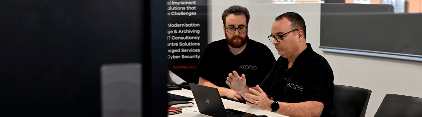 Two men discussing work at a desk with laptops in a modern office, both wearing black t-shirts with a company logo.