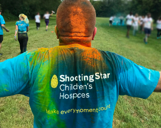A man wearing a colorful tie-dye shirt with "shooting star children's hospices - make every moment count" on the back, looking towards a group of runners in a park.