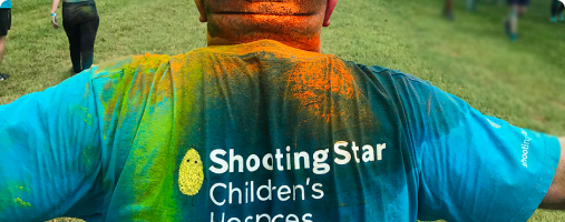 Person wearing a colorful shirt with "shooting star children's hospices" logo, covered in vibrant powder at a festive event.
