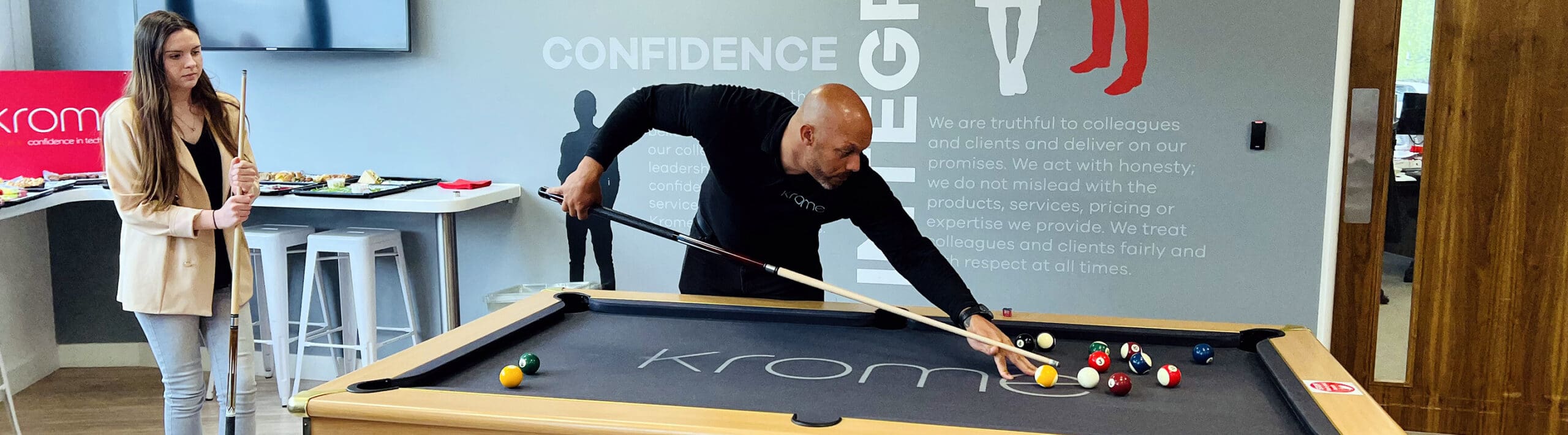 A man and woman play pool in an office game room with a company's core values displayed on the wall behind them.