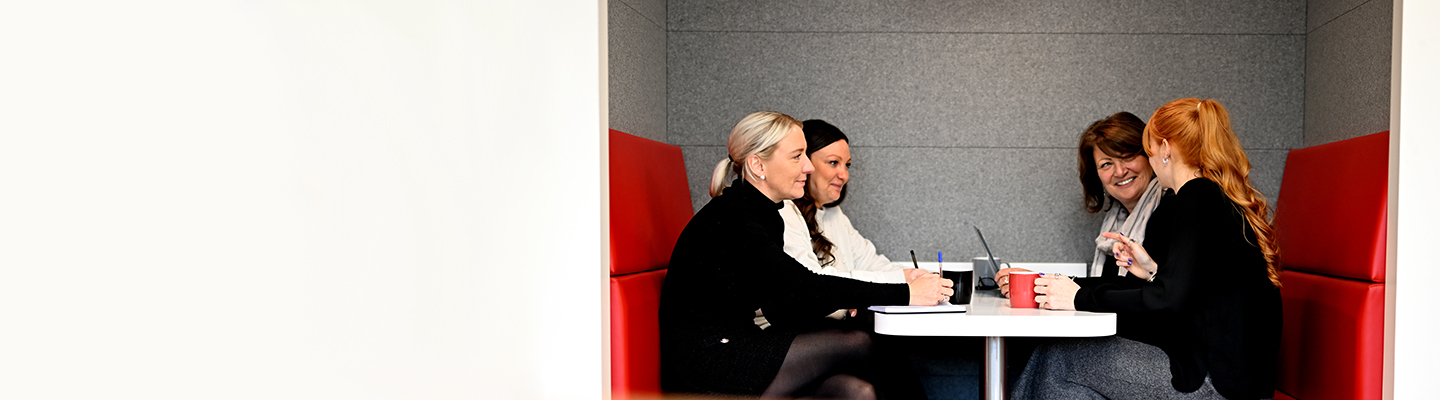 Four women sitting and laughing around a table in a modern office setting with red and grey walls.