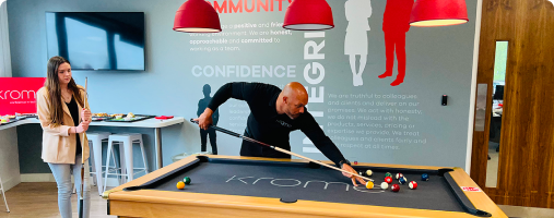 A man playing pool with a woman watching in an office game room under large red lamps, next to a wall with inspirational words.