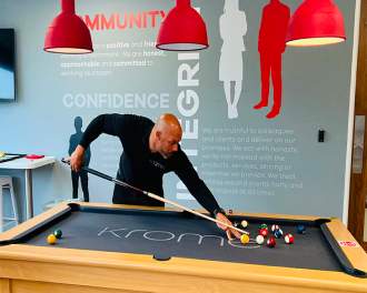 A man playing pool in an office setting, with red pendant lights overhead and values printed on the wall behind.