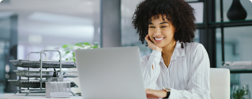A smiling woman with curly hair working on a laptop in a modern office setting.
