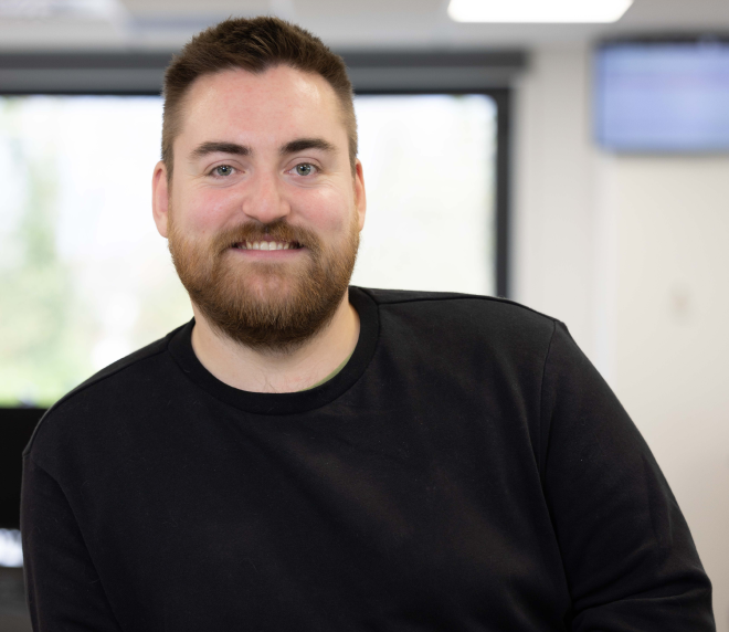 Man with a beard smiling at the camera, wearing a black shirt, in the bright Krome Team office setting.
