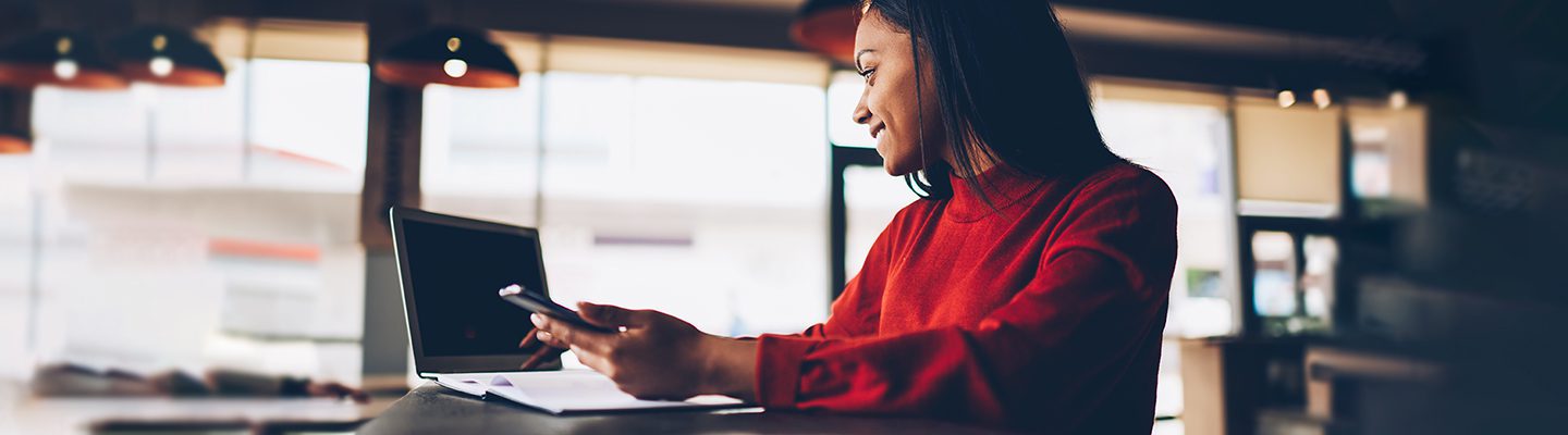 A woman in a red sweater smiles while using a smartphone and laptop at a cafe table.