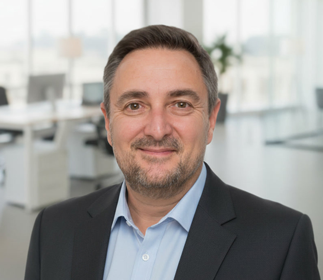 A man in a suit and light blue shirt smiles whilst standing in a modern office with large windows and desks in the background, proudly representing the Krome Team.