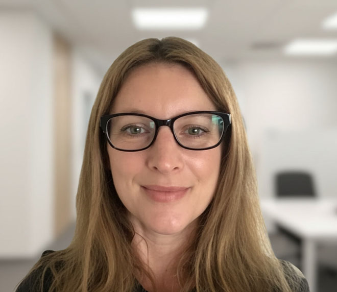 A woman with long light brown hair and glasses smiles at the camera in a bright office setting, representing the Krome Team, with white walls and blurred furniture in the background.