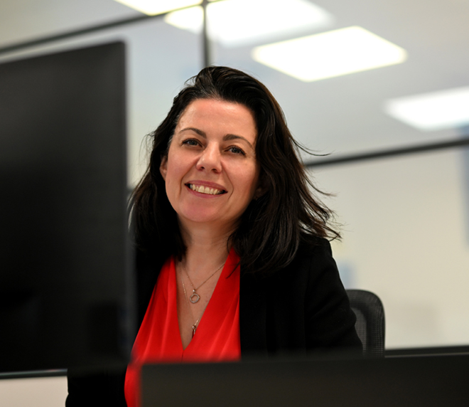 Woman with dark hair, wearing a red blouse and black blazer, smiles whilst sitting at a desk in a modern office environment.